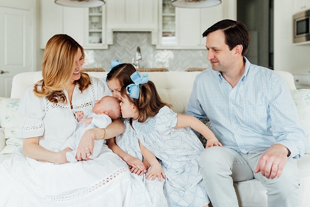 Family in living room with big sisters kissing newborn baby brother.